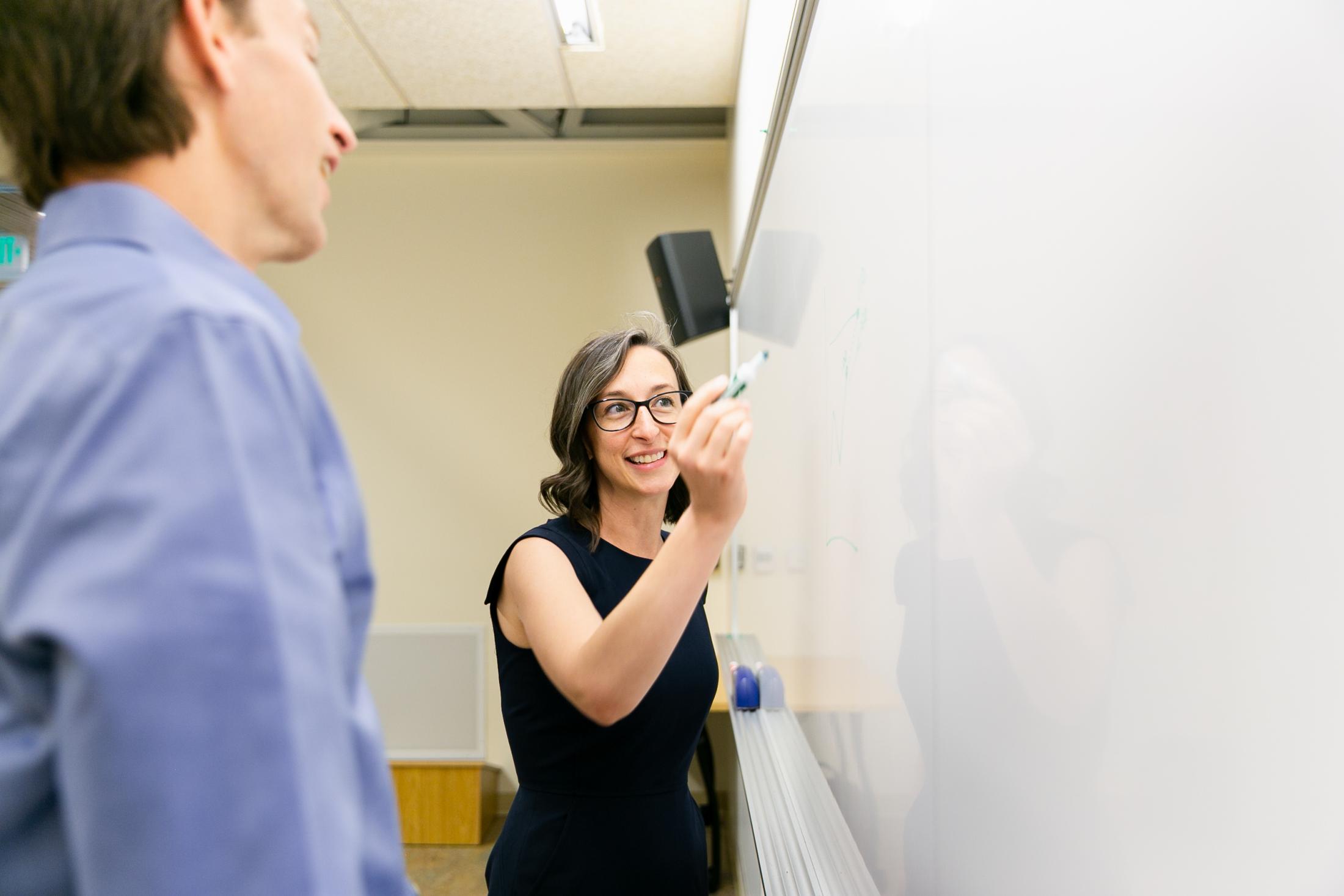 staff member writing on whiteboard and looking at colleague. 