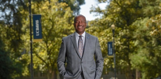 Chancellor May smiles at the camera while standing on the UC Davis Campus in front of green trees and school banners.