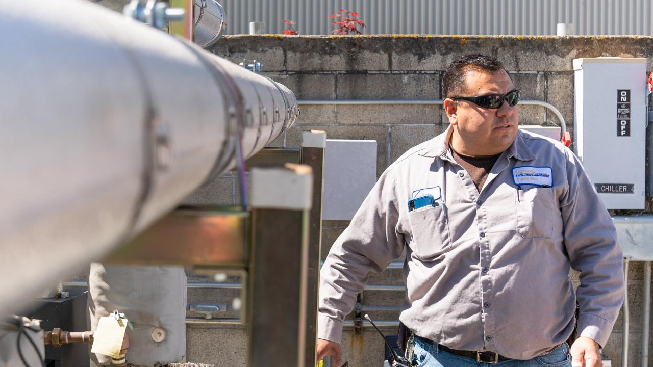 A man in a gray shirt and blue jeans walks alongside a large pipe.