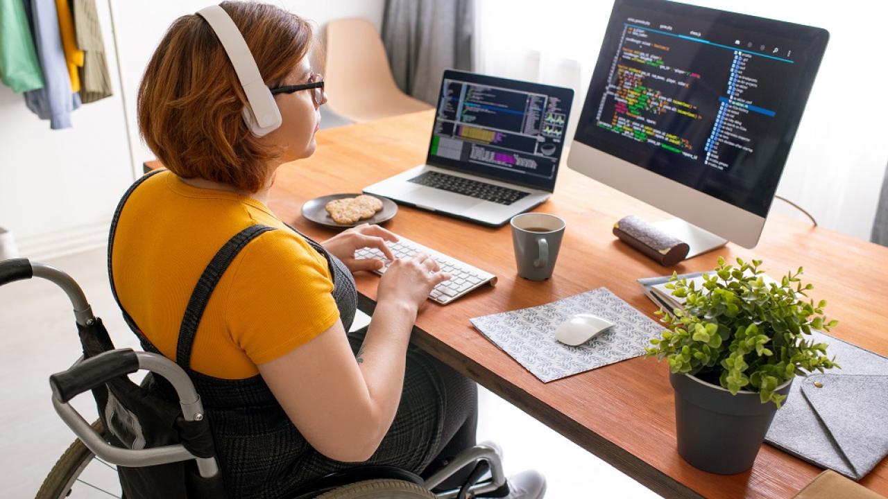 Person sitting in wheelchair at a desk, looking at a computer monitor and wearing headphones. 