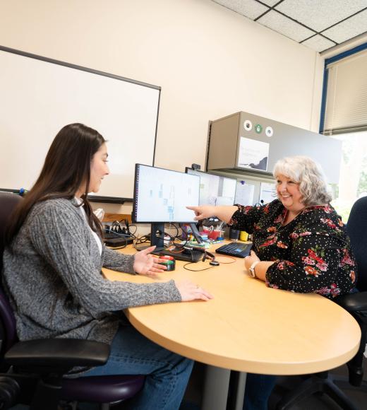 Two women sitting at a table and talking. There is a computer screen with a calendar between them that one woman points to.