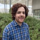 Smiling man with curly brown hair in blue plaid shirt, greenhouse photograph