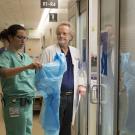 Man and woman are preparing to enter patient's room.  Woman is putting on protective gear.  