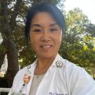 A healthcare professional in a white lab coat, smiling outdoors with greenery in the background.