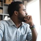 A man sitting at a desk with his laptop stares stoically out of the window while resting his head on his hand.