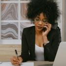 A woman with curly hair in a blazer talks on the phone while writing at a desk with a laptop.